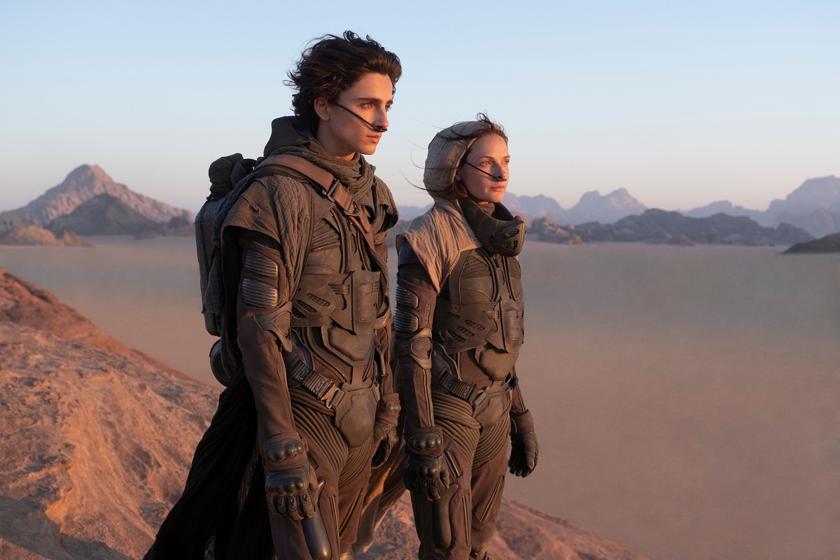Film still cu Timothee Chalamet și Rebecca Ferguson, din filmul Dune, 2021. Ei sunt îmbrăcați în armuri negre, cu un fundal cu deșert