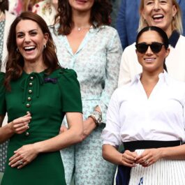 Kate Middleton și Meghan Markle, tribuna regală la Turneul de la Wimbledon, 2019. Kate poartă o rochie verde, Meghan Poartă o cămașă albă și o fustă cu o fundă neagră în talie, plus ochelari de soare
