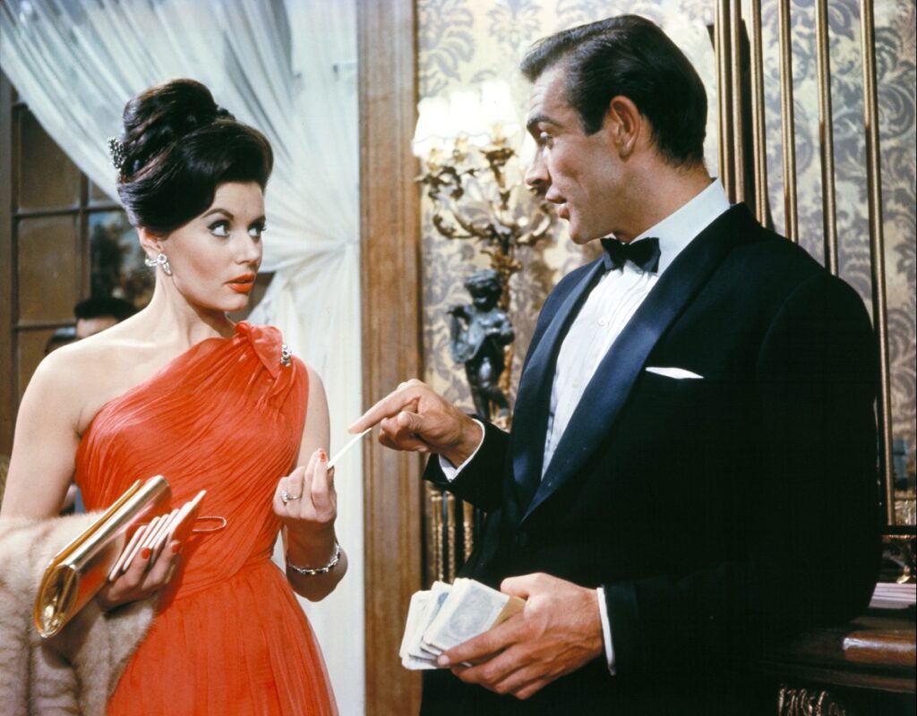 Sean Connery într-o imagine din Dr. No, primul film James Bond. Poartă un costum negru, bluză albă, și o femeie poartă o rochie roșie