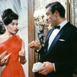 Sean Connery într-o imagine din Dr. No, primul film James Bond. Poartă un costum negru, bluză albă, și o femeie poartă o rochie roșie
