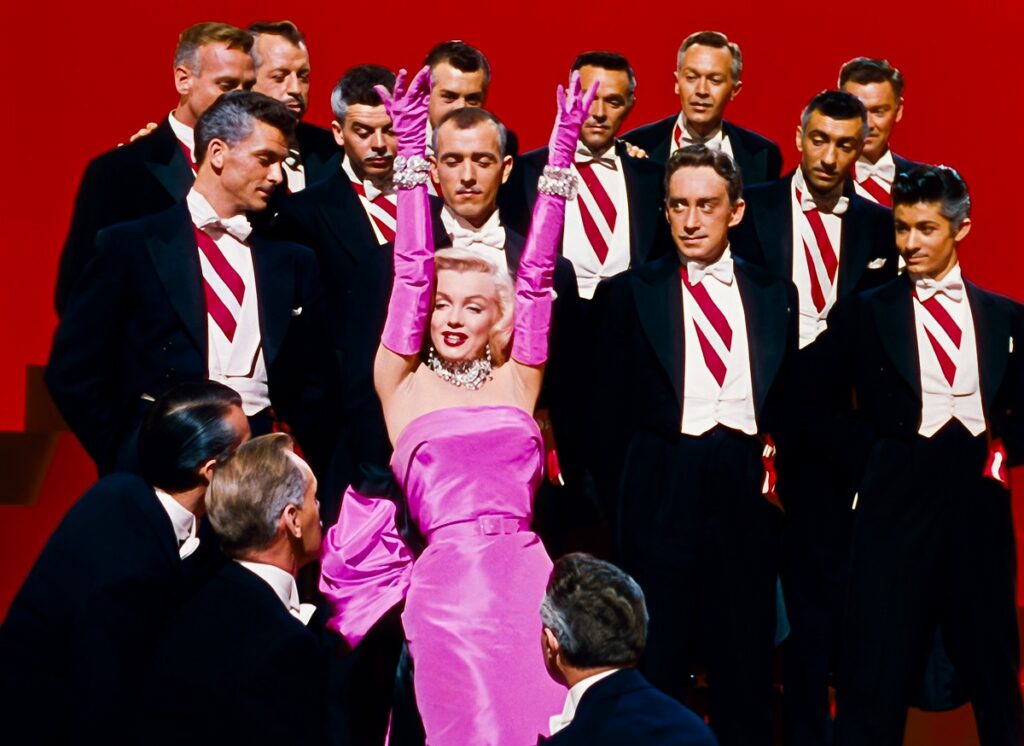 Marilyn Monroe în filmul Gentlemen Prefer Blondes. Poartă faimoasa rochie roz, înconjurată de bărbați în costume negre, cu cămpși albe, fundal roșu