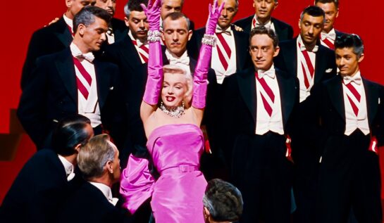 Marilyn Monroe în filmul Gentlemen Prefer Blondes. Poartă faimoasa rochie roz, înconjurată de bărbați în costume negre, cu cămpși albe, fundal roșu