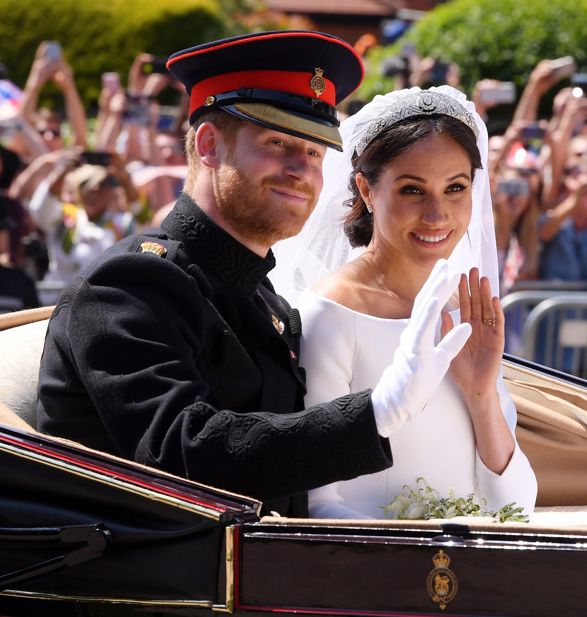 Prințul Harry și Meghan Markle la nunta lor, în trăsura neagră. Ea poartă o rochie de mireasă și văl, el poartă uniforma neagră, cu accente roșii. Fundal cu mulțime