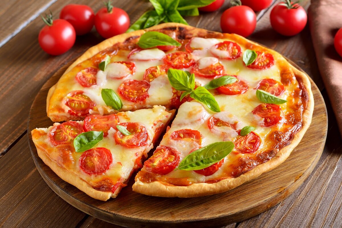 Pizza Margherita secționată pe un tocător de lemn, alături de roșii cherry și frunze de busuioc