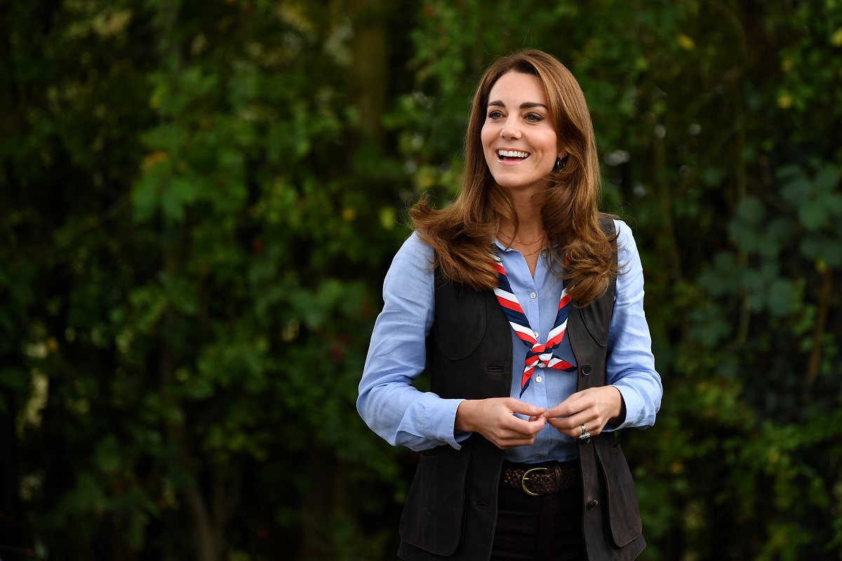 Kate Middleton în anul 2020, în timpul unui eveniment oficial din Londra. În natură, cu verdeață pe fundal, e îmbrăcată în uniformă de cercetaș, cu o cămașă albastră, vestă neagră și eșarfă la gât cu roșu
