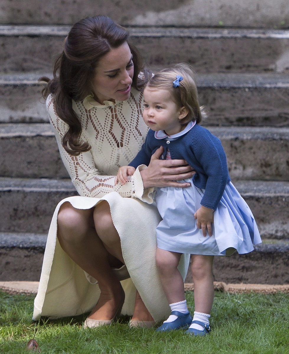 Kate Middleton și Prințesa Charlotte, împreună în Victoria, Canada, în anul 2016. Charlotte poartă o rochie albastră, Kate Middleton e îmbrăcată în bej. Ambele stau pe iarbă