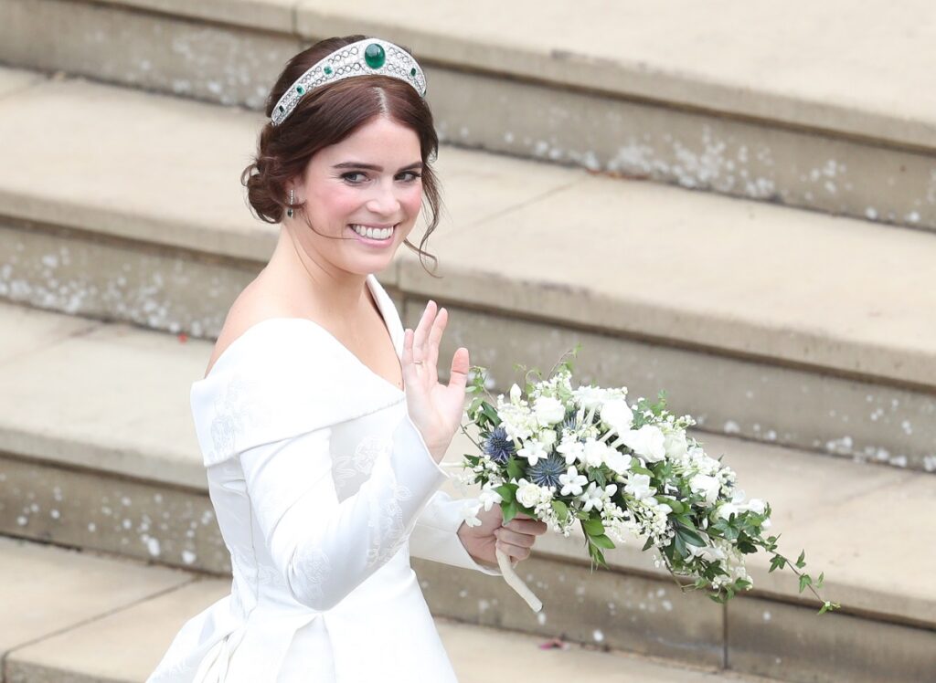 Prințesa Eugenie la nunta ei din 2018. E îmbrăcată în alb și poartă un buchet de flori albe în mână. Pe fundal scări
