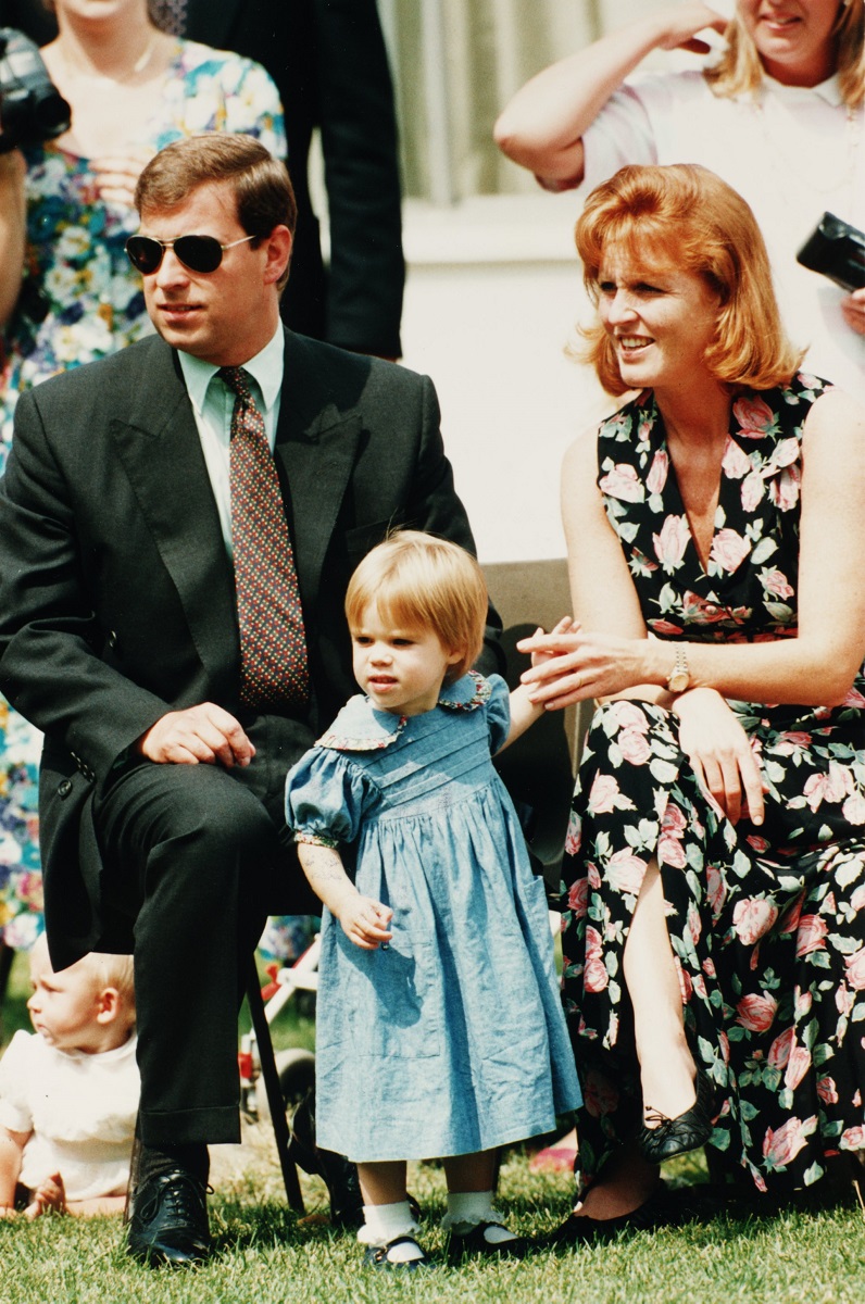 Prințul Andrew, Sarah Ferguson, Prințesa Eugenie la un eveniment oficial, pe iarbă. Sarah poartă o rochie neagră, cu motive florale, Andrew oartă costum și cravată și Eugenie poartă o rochie albastră