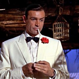 Sean Connery în rolul lui James Bond din Goldfinger. E îmbrăcat într=un costum alb, cu o floare roșie în jachetă