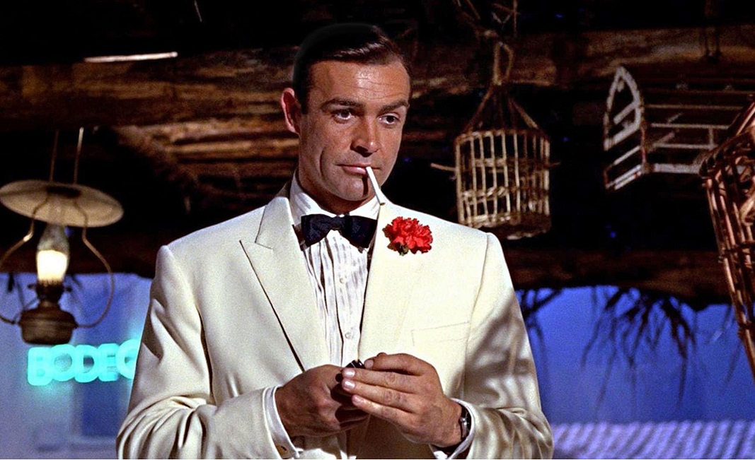 Sean Connery în rolul lui James Bond din Goldfinger. E îmbrăcat într=un costum alb, cu o floare roșie în jachetă