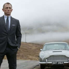 Daniel Craig în filmul Skyfall, lansat în anul 2012. Poartă un costum gri, fundal cu dealurile din Scoția, cu mașina gri în spate