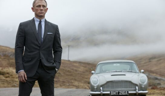 Daniel Craig în filmul Skyfall, lansat în anul 2012. Poartă un costum gri, fundal cu dealurile din Scoția, cu mașina gri în spate