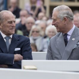 Prințul Philip și Prințul Charles la dezvelirea unei statui a Reginei Elisabeta, în 2016. Cei doi râd și se uită unul la celălalt. Philip poartă un costum albastru închis, Charles poartă un costum gri deschis