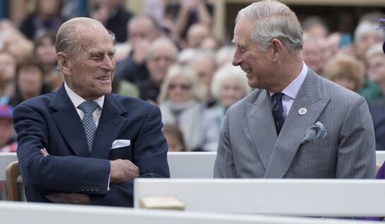 Prințul Philip și Prințul Charles la dezvelirea unei statui a Reginei Elisabeta, în 2016. Cei doi râd și se uită unul la celălalt. Philip poartă un costum albastru închis, Charles poartă un costum gri deschis