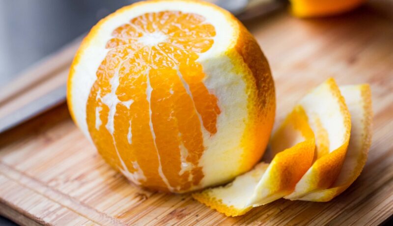 O portocală pe jumătate decojită pe un tocător de lemn în timp ce sugerează beneficiile cojilor de portocale