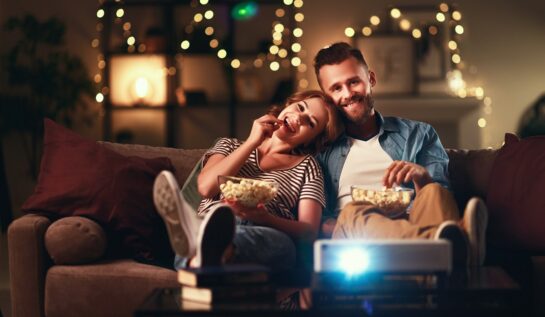 Un cuplu de îndrăgostiți care stau pe canapea și mănâncă floricele în timp ce privesc cele mai bune filme de drgoste din toate timpurile