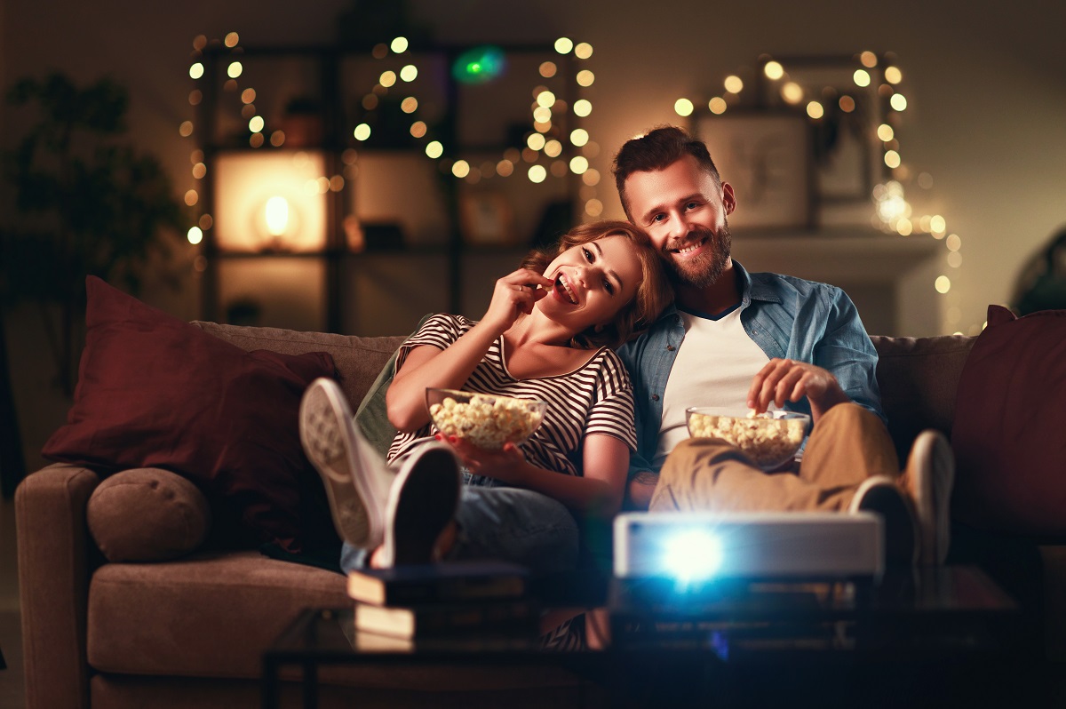 Un cuplu de îndrăgostiți care stau pe canapea și mănâncă floricele în timp ce privesc cele mai bune filme de drgoste din toate timpurile