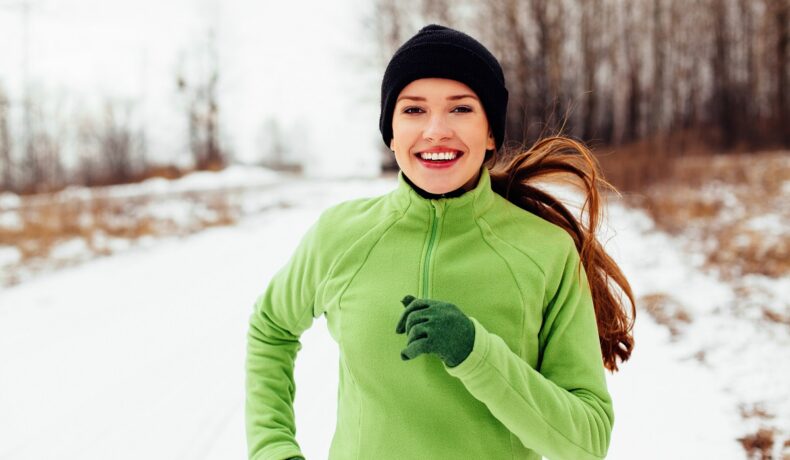 O femeie într-un hanorac verde cu o căciulă neagră pe cap în timp ce aleargă deaorece iarna este cel mai bun moment pentru a pierde în greutate