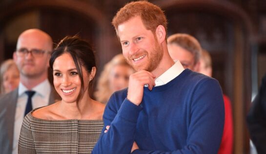 Meghan Markle într-un costum gri alături de Prințul Harry într-un pulover albastru la o ceremonie oficială din Marea Britanie