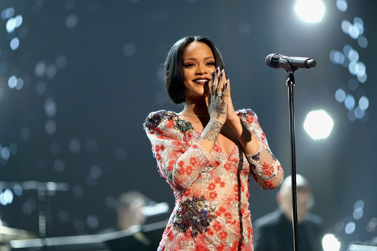 Cântăreața Rihanna într-o ținută de scenă crem cu roșu în timp ce ăși ține mâinile împreunate și zâmbește în fața unui microfon