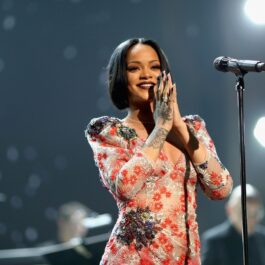 Cântăreața Rihanna într-o ținută de scenă crem cu roșu în timp ce ăși ține mâinile împreunate și zâmbește în fața unui microfon
