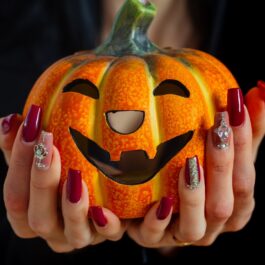 Un dovleac portocaliu cu o față zâmbitoare în timp ce este ținut în aer de două mâini cu modele de unghii pentru Halloween