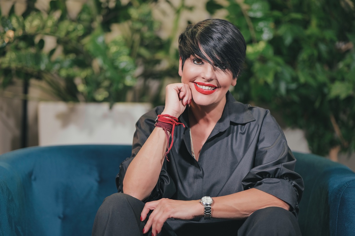 Patrizia Paglieri cu mâna sub bărbie într-o cămașă neagră, stând pe o canapea albastră la interviul CaTine. ro despre experineța Asia Express