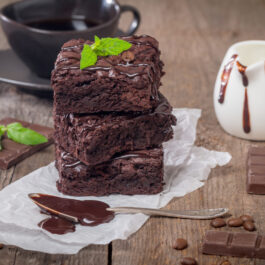 Trei bucăți de brownie, negresă americană, așezate pe un șervețel alb din hârtie, alături de bucăți de ciocolată și o spatulă cu ciocolată topită.