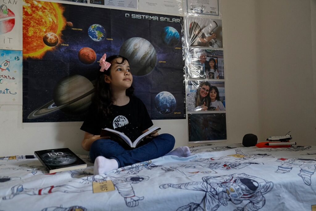 Nicole Oliveira, cea mai tânără astronomă din lume, în camera ei. Stă pe pat, are o carte în mână, în spate are poster cu planete