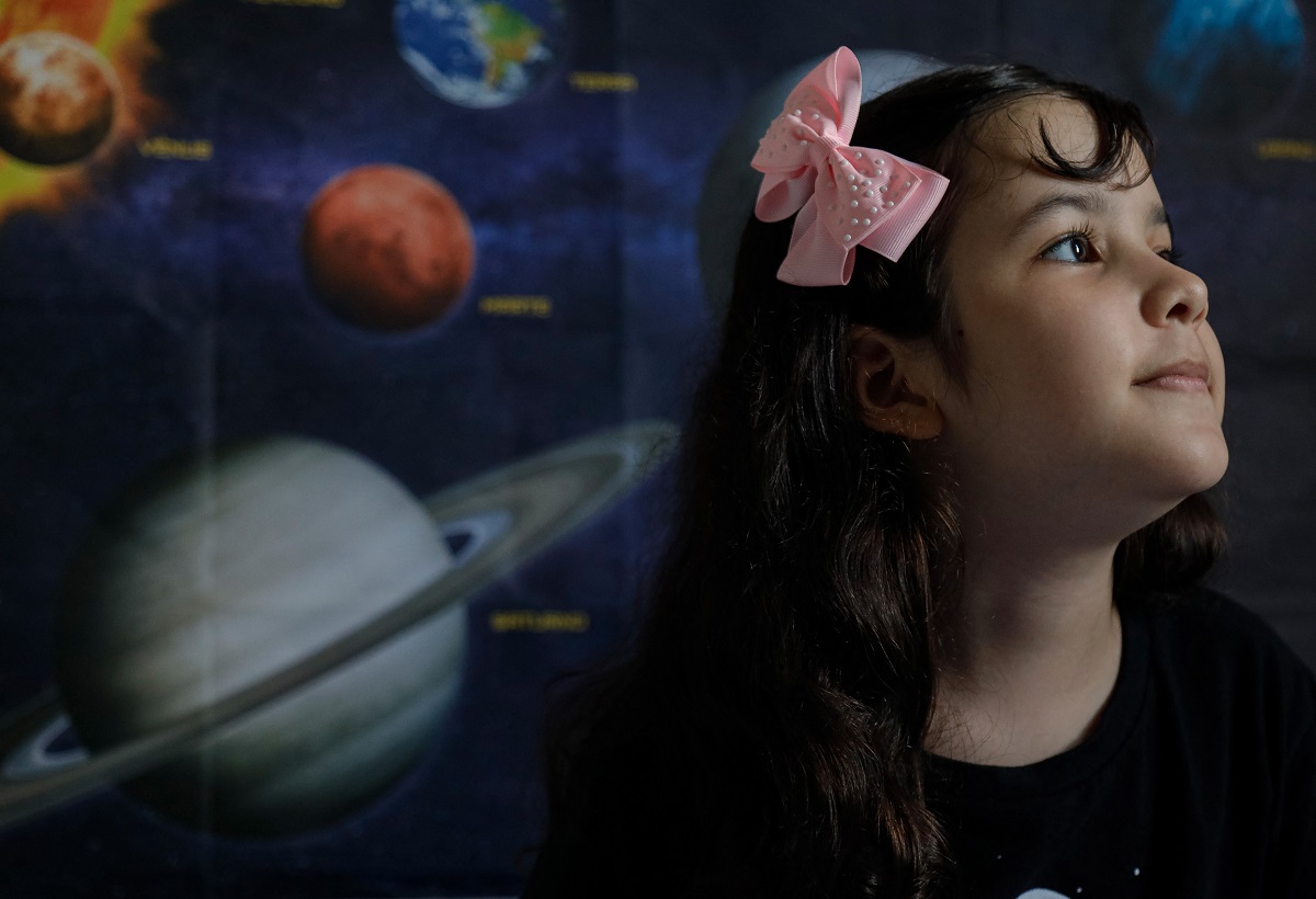 Nicole Oliveira, cea mai tânără astronomă din lume, cu o imagine cu Sistemul Solar pe fundal. Are o bluză neagră și o fundă roz în păr