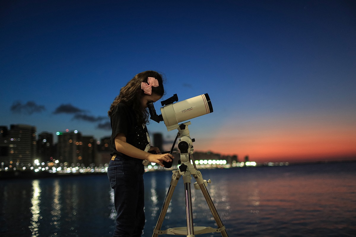 Nicole Oliveira studiază cerul cu telescopul ei alb. Pe fundal e apusul, peste un lac. Ea e îmbrăcată în negru