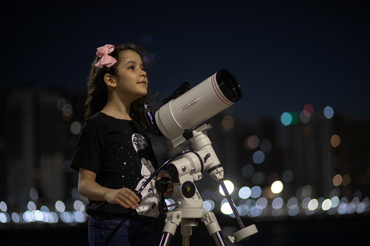 Nicole Oliveira, cea mai tânără astronomă din lume, studiază cerul cu telescopul alb. E îmbrăcată în negru, pe fundal e un oraș