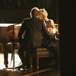 Lady Gaga pe scenă alături de Bradley Cooper care o ține în brațe în timp ce cântă împreună la pian la Premiile Oscar din 2019