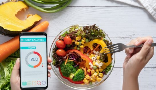 o masă pe care se află un bol cu salată în timp ce o femeie ține o fruculiță într-o mână iar în cealaltă un telefon pe care numără caloriile recomandate zilnic