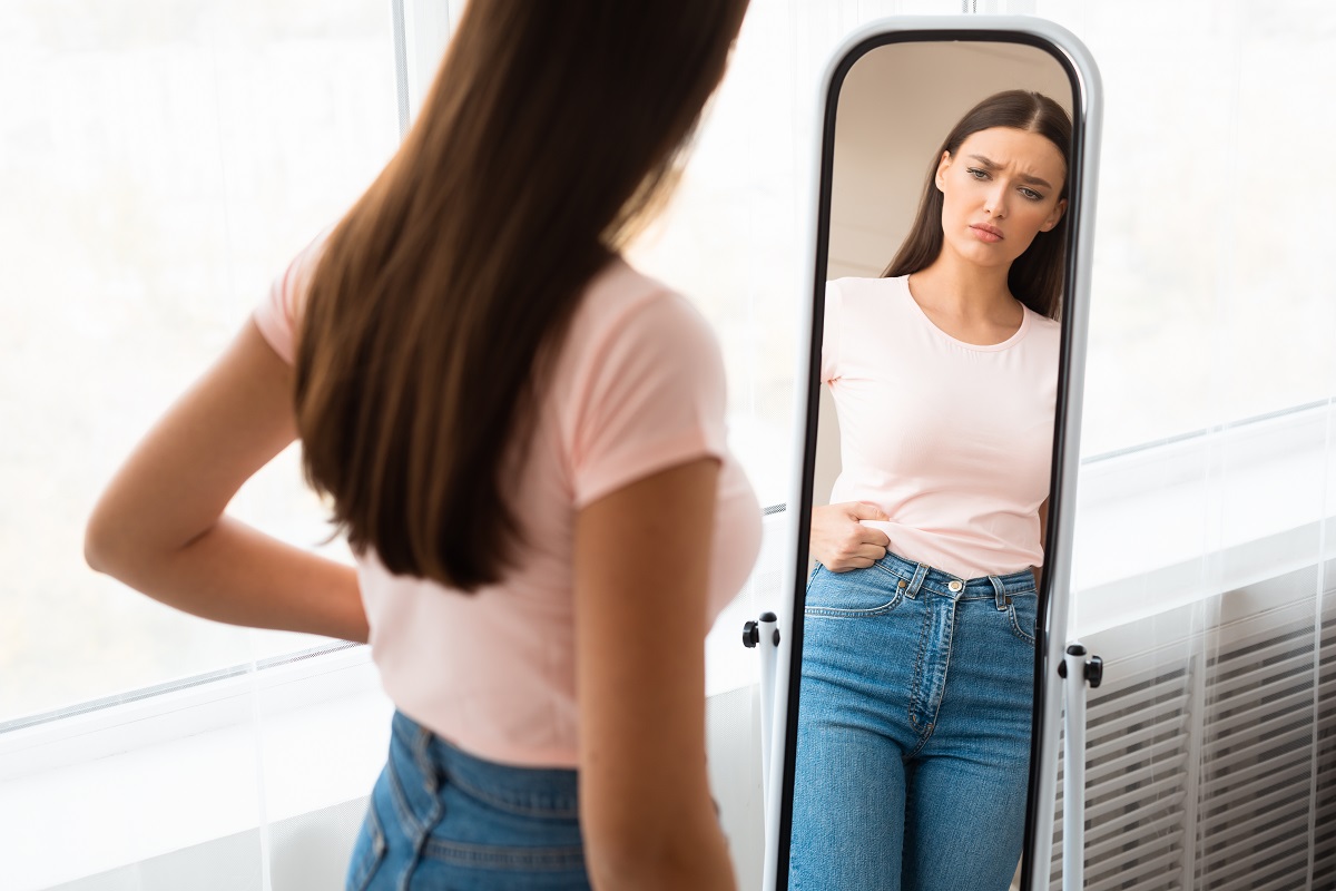 O femeie frumoasă care stă cu fața la oglindă în timp ce privește la grăsimea abdominală pe care o are