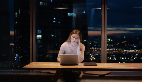 O femeie blondă care stă în fața unui laptop la birou în timp ce își sprijină capul în mâini, afară fiind noapte, care simbolizează starea sa afectată de lipsa somnului