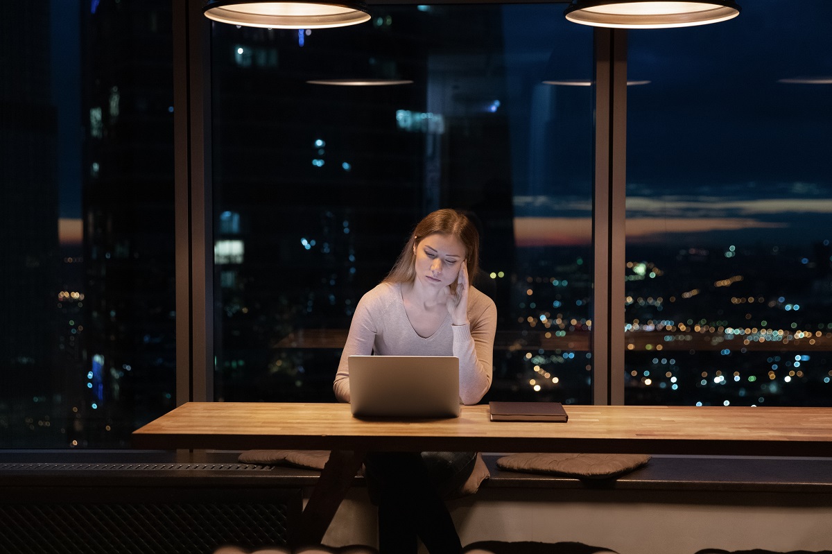 O femeie blondă care stă în fața unui laptop la birou în timp ce își sprijină capul în mâini, afară fiind noapte, care simbolizează starea sa afectată de lipsa somnului