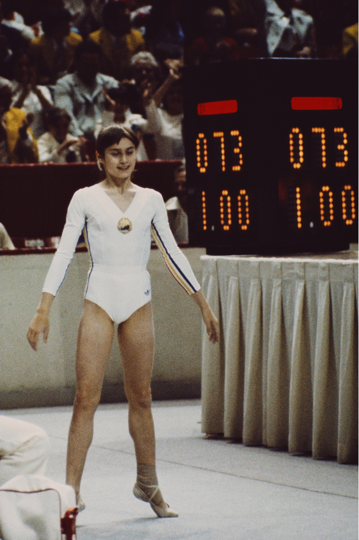 Nadia Comăneci într-un costum alb de gimnastică perdormând la sol la Jocurile Olimpice de vară din 1976