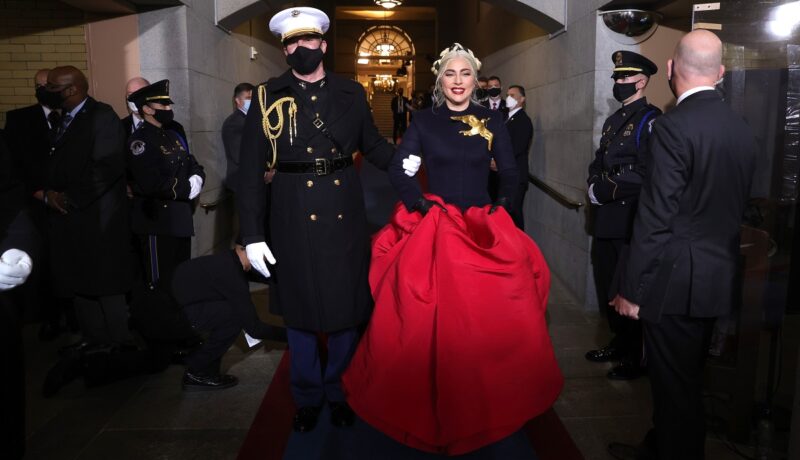 Lady Gaga într-o rochie neagră cu o trenă roșie la brațele unui soldat în timp ce merge să cânte imnul național al SUA la inaugurarea președintelui Joe Biden