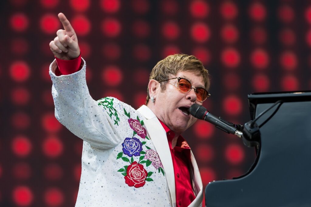 Sir Elton John într-un sacoul alb și o cămașă albă în timp ce cântă la pian la un concert fiind una din celebritățile care și-au schimbat numele