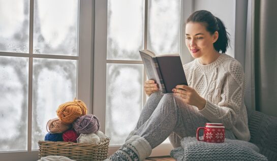 O femeie frumoasă îmbrăcată în haine călduroase de iarnă în timp ce stă la fereastră și citește autori români contemporani
