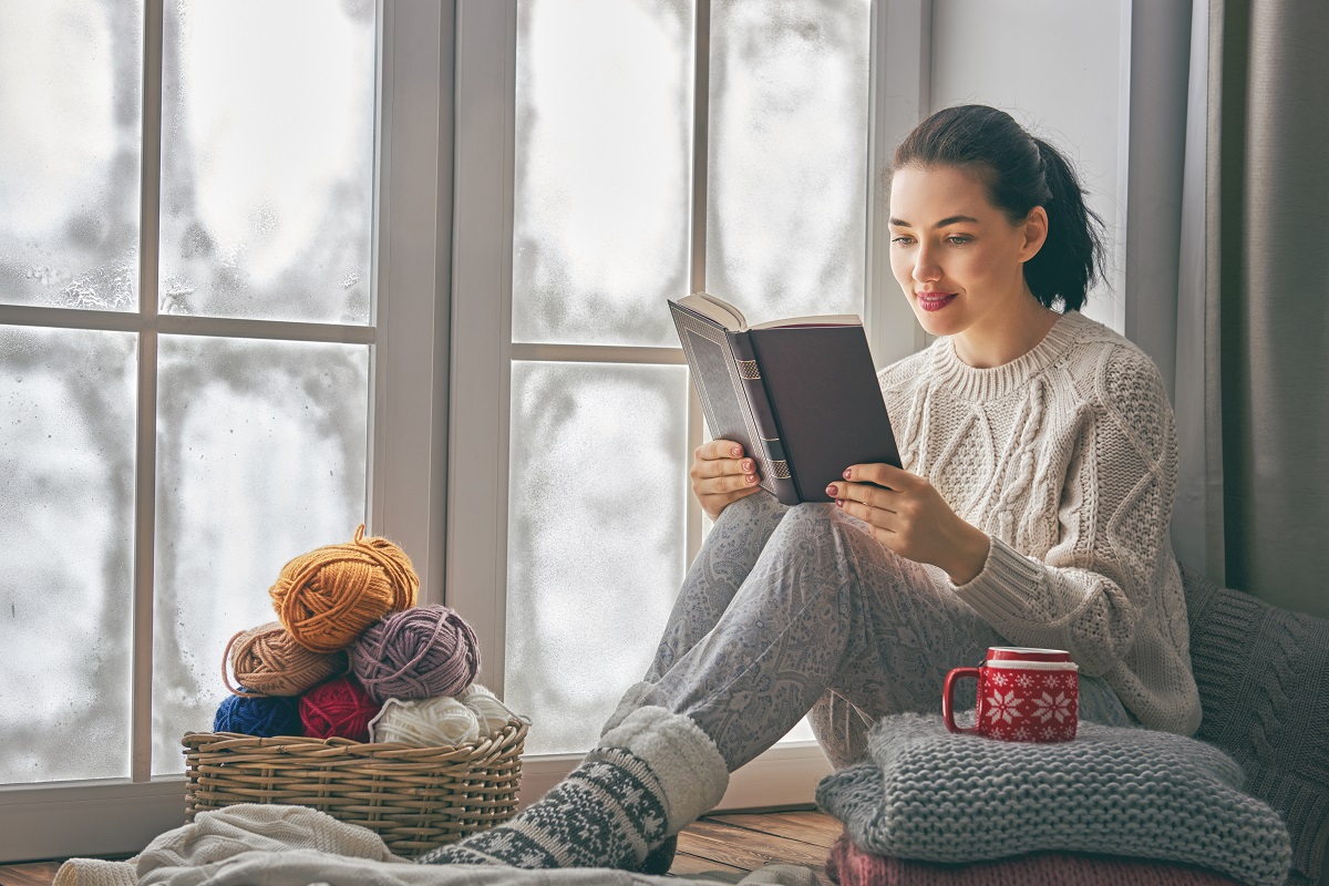 O femeie frumoasă îmbrăcată în haine călduroase de iarnă în timp ce stă la fereastră și citește autori români contemporani