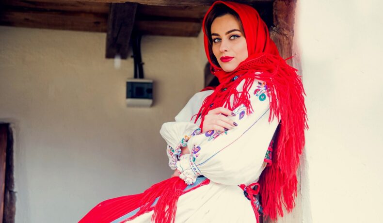 O femeie frumoasă îmbrăcată în port popular românesc în timp ce stă pe prispa unei case cu o basma roșie pe cap