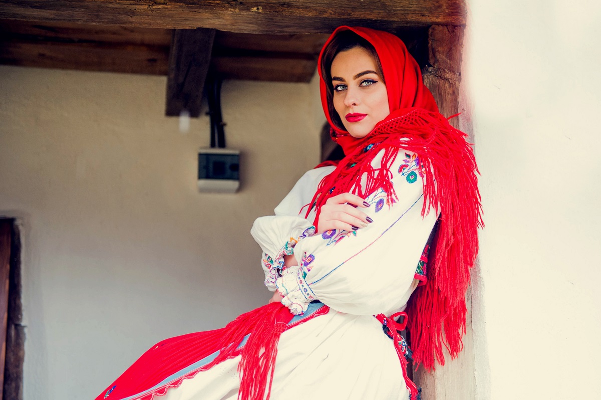 O femeie frumoasă îmbrăcată în port popular românesc în timp ce stă pe prispa unei case cu o basma roșie pe cap