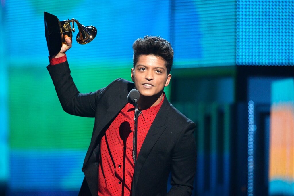 Bruno Mars la costum negru cu o cămașă roșie ridicând un trofeu Grammy