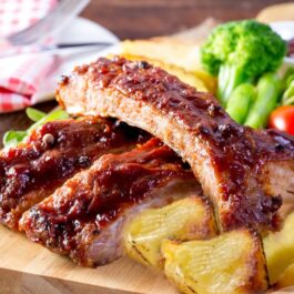 Coaste de porc cu cartofi pe un suport de lemn, alături de legume proaspete