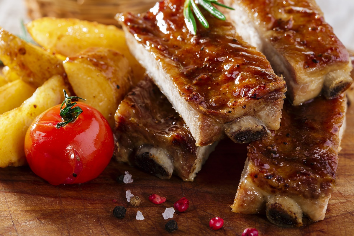 Coaste de porc cu cartofi la cuptor, pe un suport de lemn, alături de o roșie coaptă