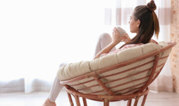 O femeie care își bea cafeaua la geam, pe un fotoliu confortabil