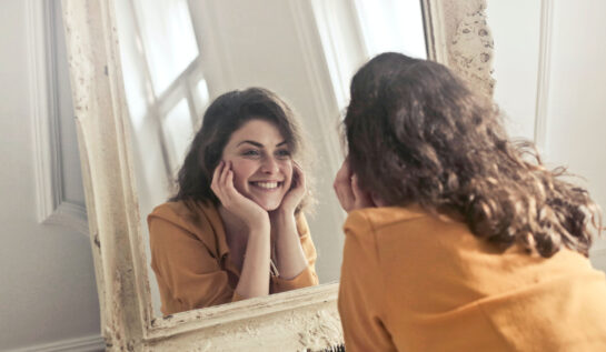 O femeie care zâmbește în oglindă, îmbrăcată cu o bluză portocalie.