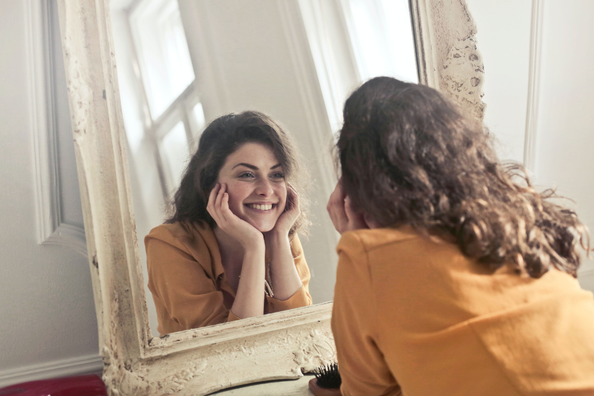 O femeie care zâmbește în oglindă, îmbrăcată cu o bluză portocalie.